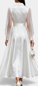 Back view of a Mahaute satin midi shirt dress in white with sheer long sleeves and a tied waist, worn with heels.