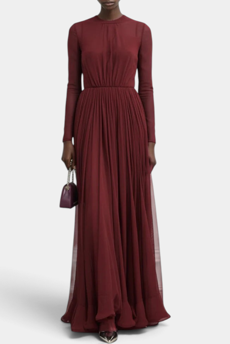 Mahaute burgundy long-sleeved pleated maxi dress with a high neckline worn by a model holding a small clutch bag