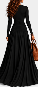 Mahaute Gracia long black dress with long sleeves, ruched waist, and flowing skirt, worn by a woman holding a bag.