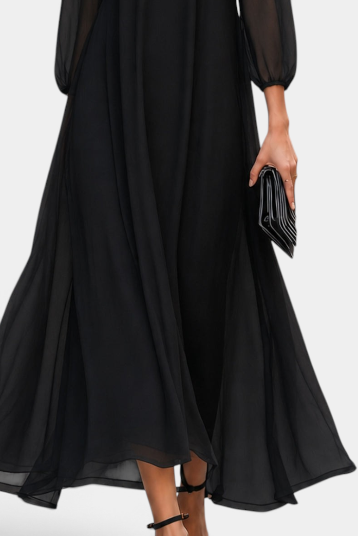 Mahaute Mazarine long black flowing dress with sheer sleeves and flared skirt, worn with elegant heels and a clutch.