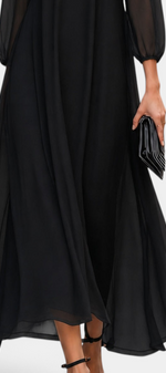 Mahaute Mazarine long black flowing dress with sheer sleeves and flared skirt, worn with elegant heels and a clutch.