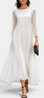 Mahaute Mazarine long white flowing dress with sheer sleeves and flared skirt, styled with black heels and a clutch.