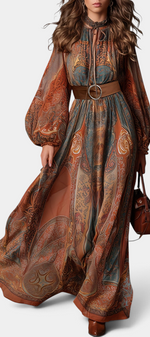 Mahaute Suevy long bohemian dress with ornamental print, tie neck, sheer puff sleeves, and a brown round-buckle belt.