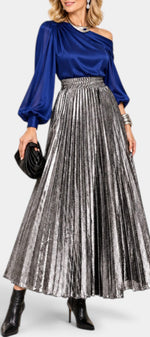 Mahaute Tegan high-waisted metallic pleated long skirt in silver, styled with a blue blouse and black ankle boots.