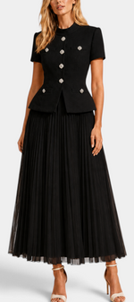Model wearing a Mahaute black structured short-sleeve jacket with jewel buttons and a matching long pleated skirt.